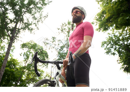 Strong Male cyclist in sportswear, glasses and protective helmet walking with his bike in the forest to take a break after riding. Sky blue and forest in the background. 91451889