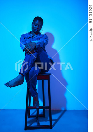 confident plus size african woman in denim shirt and trousers posing on chair in studio on blue neon background 91451924