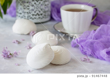 Delicate vanilla marshmallow on a white delicate plate. Still life with marshmallows, a cup of tea and lilac lilac on a background of ancient stone. Delicate vanilla marshmallow on a white delicate plate. Still life with marshmallows, a cup of tea and lilac lilac on a background of ancient stone. 91467448