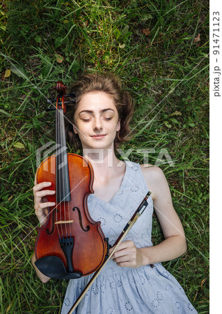 The girl lies with a violin on the grass in a city park. The girl lies with a violin on the grass in a city park. 91471123