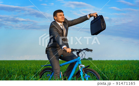 businessman rides a bicycle through a green grass field, dressed in a business suit, he has a briefcase and documents, beautiful nature in spring, business concept 91471165