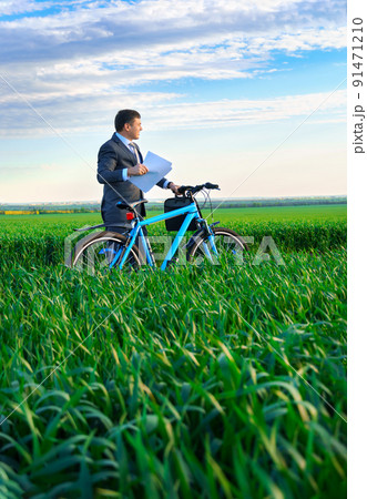 businessman dressed in a business suit, poses with bicycle in green grass field and reads documents or reports, beautiful nature in spring, business concept businessman dressed in a business suit, poses with bicycle in green grass field and reads documents or reports, beautiful nature in spring, business concept 91471210