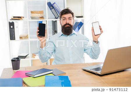 Serious businessman holding mobile phones in hands sitting at office desk, choice Serious businessman holding mobile phones in hands sitting at office desk, choice 91473112