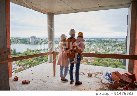 Back view of parents with daughter standing inside apartment building under construction. Man and woman with child wearing building helmets while enjoying city view from future home. Back view of parents with daughter standing inside apartment building under construction. Man and woman with child wearing building helmets while enjoying city view from future home. 91476932