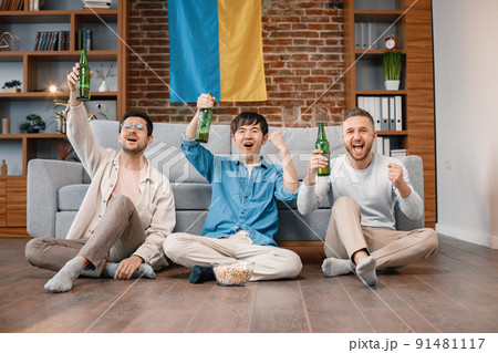 Male friends gathered at home to watch a football match. Men sitting on a floor near the couch in front of big flag of Ukraine on a wall. Men actively support their favorite team and feeling satisfied 91481117