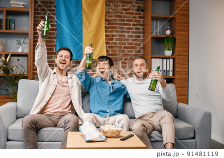 Male friends gathered at home to watch a football match. Men sitting on the couch in front of big flag of Ukraine on a wall. Men actively support their favorite team and feeling satisfied. 91481119