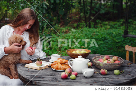 woman enjoying morning coffee in apple orchard with her pet poodle 91483008