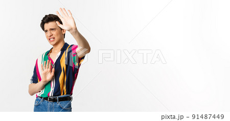 Timid and displeased androgynous man asking to stop, raising hands defensive and grimacing, standing over white background Timid and displeased androgynous man asking to stop, raising hands defensive and grimacing, standing over white background 91487449
