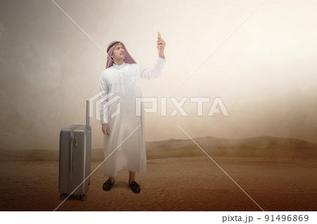 Arab man wearing keffiyeh looking at mobile phone with a suitcase for traveling 91496869