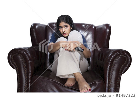Asian woman holding a television remote sitting on the couch with a scared expression Asian woman holding a television remote sitting on the couch with a scared expression 91497322