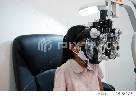Ophthalmologist examining the eyes of an Asian girl in a clinic. They wear protective face masks. 91498472