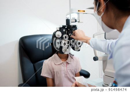 Ophthalmologist examining the eyes of an Asian girl in a clinic. They wear protective face masks. Ophthalmologist examining the eyes of an Asian girl in a clinic. They wear protective face masks. 91498474
