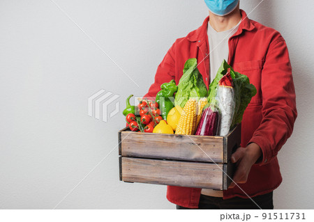 Young delivery man in protective face mask holding wooden box in hands. Man courier with shopping box. Safe online delivery from supermarket to home. COVID-19 coronavirus protection Young delivery man in protective face mask holding wooden box in hands. Man courier with shopping box. Safe online delivery from supermarket to home. COVID-19 coronavirus protection 91511731