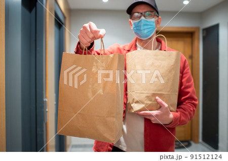 Delivery man in protective mask holding paper bag with food in the entrance. The courier gives the box with fresh vegetables and fruits to the customer Delivery man in protective mask holding paper bag with food in the entrance. The courier gives the box with fresh vegetables and fruits to the customer 91512124