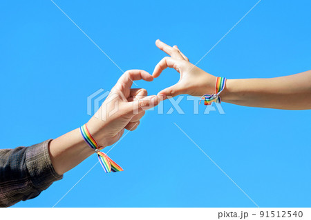 Close up of Woman hands in a rainbow bracelet making a heart shape form on blue sky background. 91512540