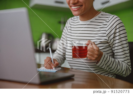 Close up shot of happy young woman holding cup of tea and writing in notepad using laptop 91520773