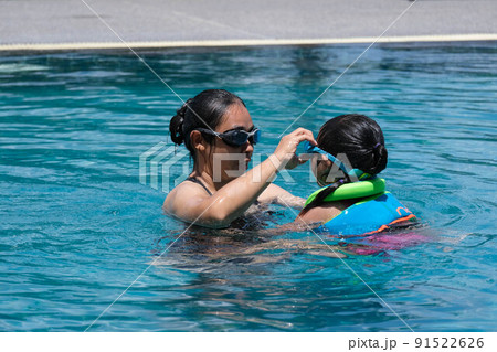 Cute little girl having fun swimming with her mother in the pool on a sunny day. Happy family, mother and her daughter playing in the swimming pool. Summer lifestyle concept. 91522626