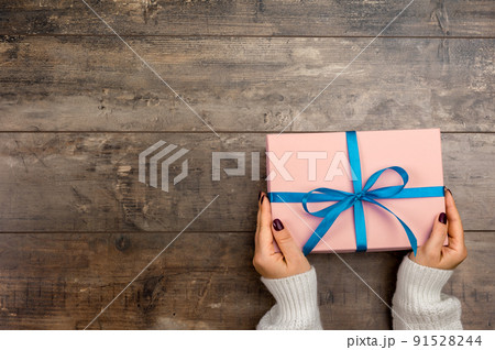 Cropped view of woman holding wrapped christmas gift with blue bow. Female hands holding gift box on wooden rustic background 91528244
