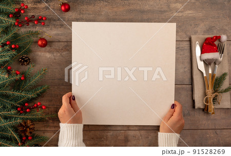 Woman hands holds menu in her hands while sitting at Christmas dinner table. Menu mockup. blank black paper on rustic wooden background. Christmas menu concept with cutlery 91528269