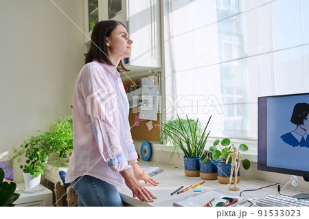 Young woman looking out the window standing near workplace in home office 91533023