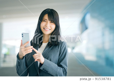 オフィスのエントランスでスマホを見る若いスーツのビジネスウーマン オフィスのエントランスでスマホを見る若いスーツのビジネスウーマン 91538781