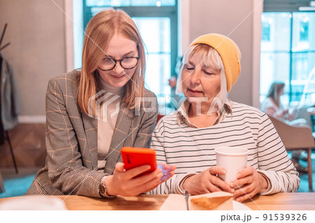 Two beautiful women friends outdoors with coffee in park using mobile phone, modern office building in the background, good for modern lifestyle. Two beautiful women friends outdoors with coffee in park using mobile phone, modern office building in the background, good for modern lifestyle. 91539326