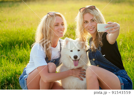 two beautiful and charming blonde twins woman with a white fluffy samoyed dog sitting and making selfie on the phone in the park two beautiful and charming blonde twins woman with a white fluffy samoyed dog sitting and making selfie on the phone in the park 91543559