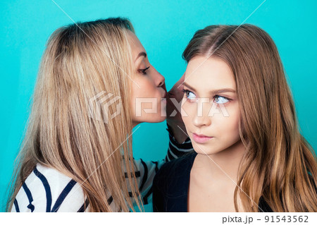 portrait of two young attractive happy women with blonde hair and makeup eavesdrops whispers a secret mystery in the studio on a blue background. the concept of gossip and confidentiality 91543562