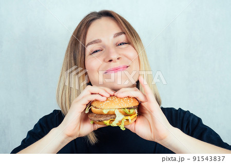 young and attractive blonde woman in black T-shirt and measuring tape enjoying a high-calorie burger. slimming and diet concept young and attractive blonde woman in black T-shirt and measuring tape enjoying a high-calorie burger. slimming and diet concept 91543837