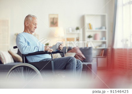 Serious concentrated senior man sitting in wheelchair and using modern app on tablet in living room Serious concentrated senior man sitting in wheelchair and using modern app on tablet in living room 91546425