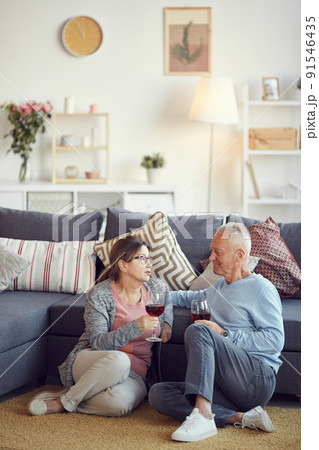 Senior brown-haired woman in glasses sitting on carpet in living room and chatting with husband while drinking wine Senior brown-haired woman in glasses sitting on carpet in living room and chatting with husband while drinking wine 91546435