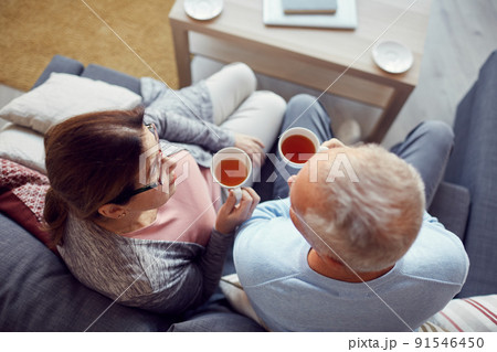 Directly above view of senior couple in casual clothing sitting on sofa in living room and drinking tea from cups while talking to each other Directly above view of senior couple in casual clothing sitting on sofa in living room and drinking tea from cups while talking to each other 91546450