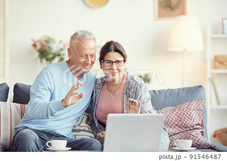Smiling seniors in casual clothing sitting on sofa in living room and waving hands while talking to grandchildren via computer app Smiling seniors in casual clothing sitting on sofa in living room and waving hands while talking to grandchildren via computer app 91546487