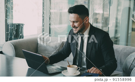Cheery bearded Caucasian businessman in formal clothes chatting to his family through laptop and waving his hand happily in a classy cafe. He gesticulates optimistically and cheerfully. 91558996