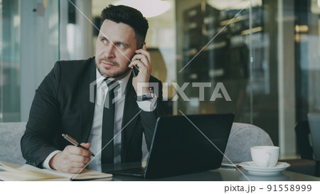 Smart Caucasian businessman sitting, talking on smartphone and writing down business information in his notepad while looking at laptop in glassy cafe 91558999