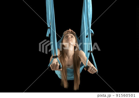 Young woman gymnast hovers in the air in sports hammock. Portrait of woman during yoga training on black background. Young woman gymnast hovers in the air in sports hammock. Portrait of woman during yoga training on black background. 91560501