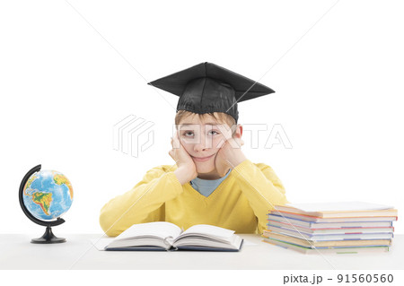 Portrait of bored schoolboy in students hat at table next to notebooks and globe. Boy sitting at his desk. Isolated on white background. Portrait of bored schoolboy in students hat at table next to notebooks and globe. Boy sitting at his desk. Isolated on white background. 91560560
