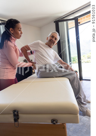 Biracial female physiotherapist giving back massage therapy to caucasian senior man at home 91561033