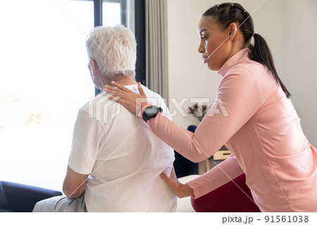 Biracial female physiotherapist giving back massage therapy to caucasian senior man at home 91561038