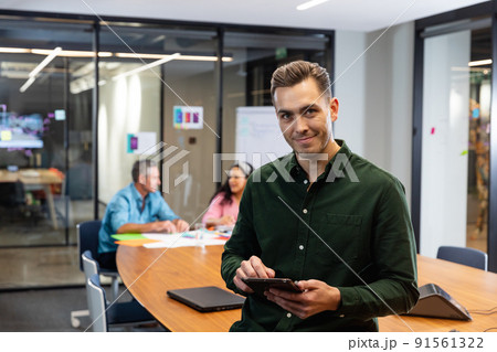 Portrait of confident young caucasian businessman with tablet pc while colleagues in background Portrait of confident young caucasian businessman with tablet pc while colleagues in background 91561322