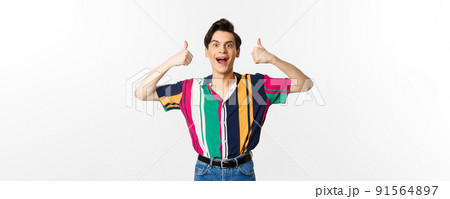 Excited and amazed young man showing thumbs up with impressed expression, standing over white background 91564897
