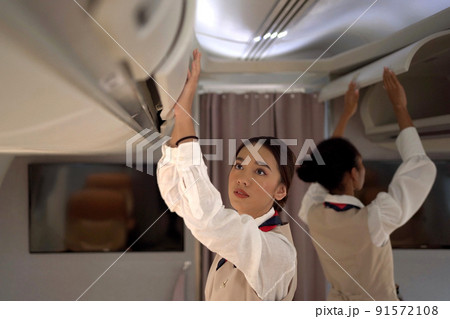Two woman cabin crews closing the overhead compartment for safety and security check before flight departure. Female flight attendant checking the cabin overhead compartment for safety instruction. Two woman cabin crews closing the overhead compartment for safety and security check before flight departure. Female flight attendant checking the cabin overhead compartment for safety instruction. 91572108