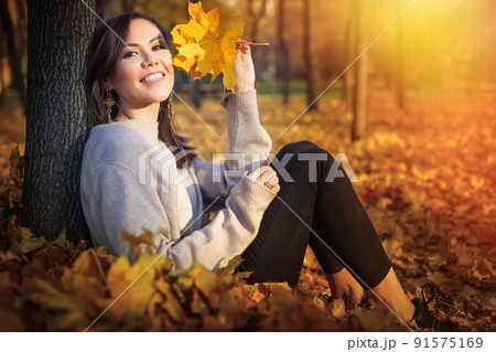 Young happy Central Asian woman sitting near tree and smiling at sunset in autumn park Young happy Central Asian woman sitting near tree and smiling at sunset in autumn park 91575169