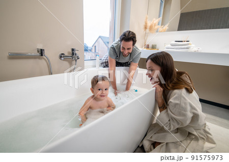 Child looking at camera in bathtub and parents watching 91575973