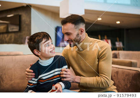 Handsome young dad communicating with his adorable son, sitting together on a comfortable couch in the VIP lounge of the airport departure terminal 91579493