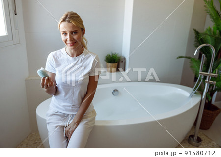 Beautiful young blonde happy woman sitting in bathroom and holding brush cleanser.  91580712