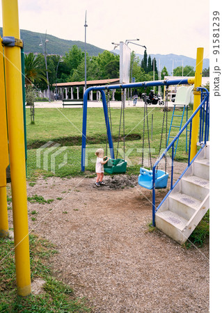 Little girl holds on to the swing with her hands on the background of buildings and park Little girl holds on to the swing with her hands on the background of buildings and park 91581239