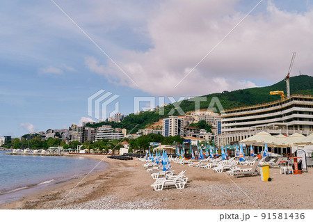 Sun loungers with folded umbrellas stand on the beach against the backdrop of buildings, greenery and mountains Sun loungers with folded umbrellas stand on the beach against the backdrop of buildings, greenery and mountains 91581436