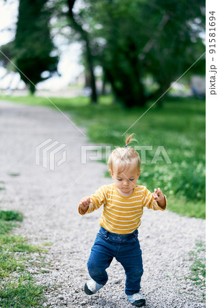Little girl walks along the gravel path in the park and looks under her feet Little girl walks along the gravel path in the park and looks under her feet 91581694