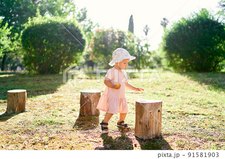 Little girl in a hat stands on the lawn near the stumps 91581903